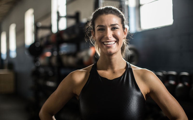 A white-presenting woman in a black tank top grins in a gym.