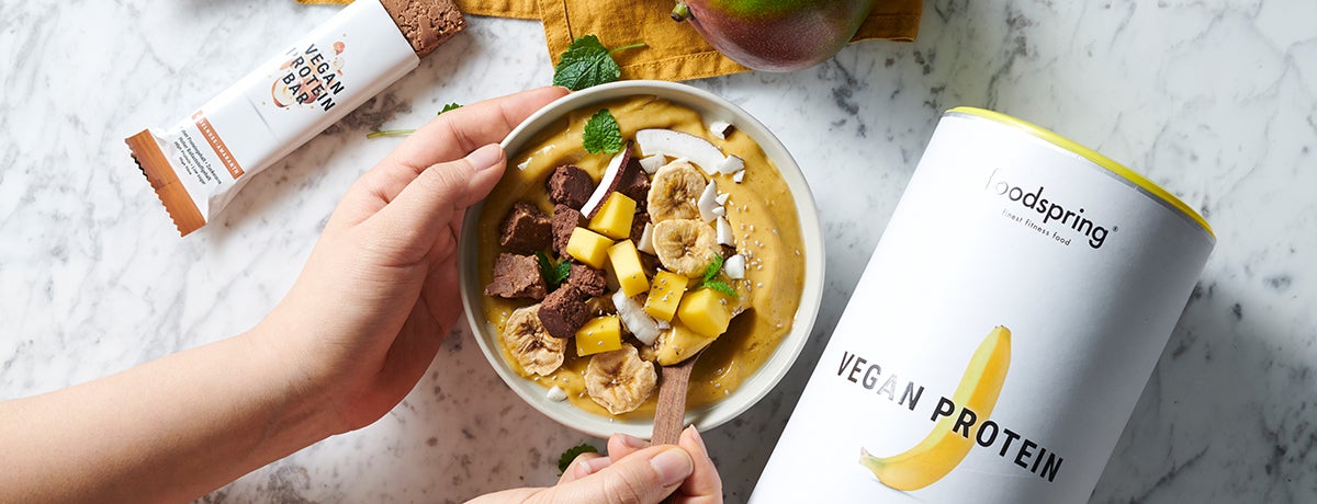 A pair of white-presenting hands holds a white stoneware bowl filled with a vitamin D rich, yellow, vegan protein smoothie bowl topped with banana chips, mango cubes, coconut chips, and vegan protein bar chunks