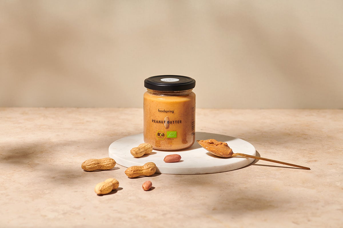 a jar of peanut butter on a white marble plate, surrounded by shelled and unshelled peanuts