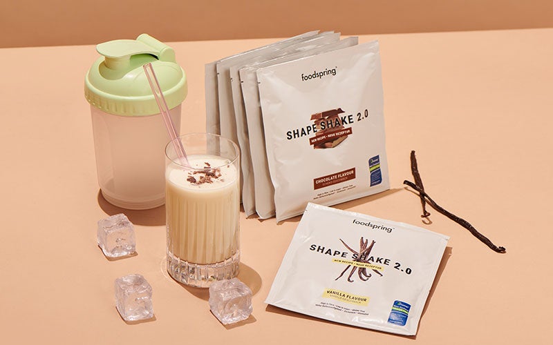 A stack of single-serving packets of Shape Shake 2.0 meal replacement shakes stand next to a plastic shaker with a green lid and a glass filled with a prepared Shape Shake 2.0 meal replacement shake garnished with a pinch of cinnamon. A few ice cubes stand on one side and two vanilla beans on the other side.