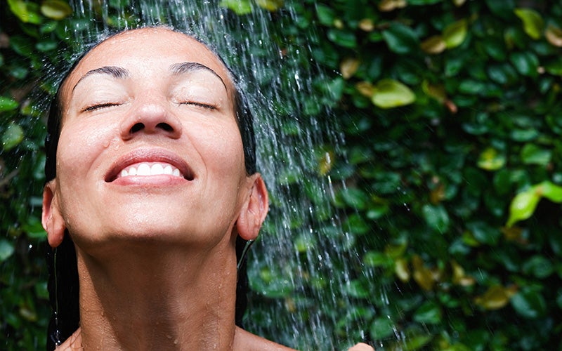 A dark-haired white woman takes a shower in front of a backdrop of leaves. Only her head and neck are visible. It seems she is showering outside.