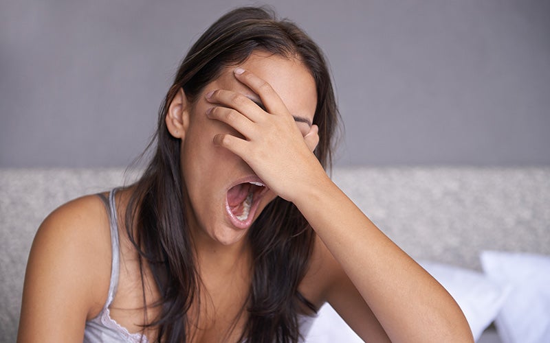 A woman with medium skin and straight brown hair covers her eyes and yawns