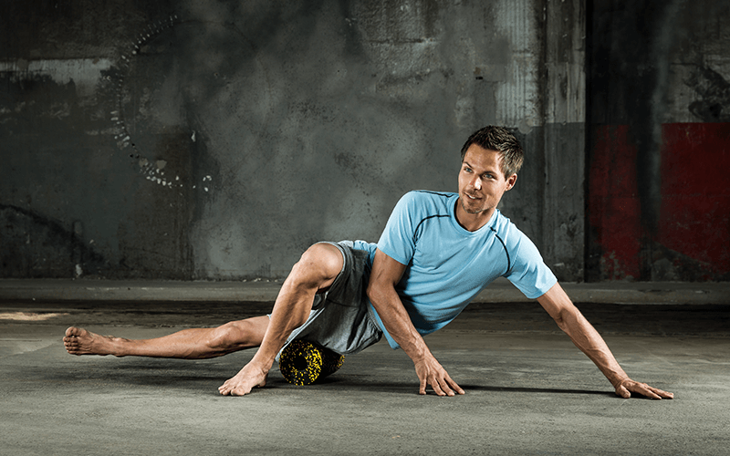 A white man in fitness clothing performs foam roller exercises by rolling his outer thigh over a foam roller