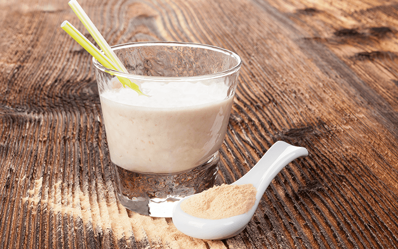 A glass of white smoothie sits next to a white spoonful of maca powder on a wooden surface