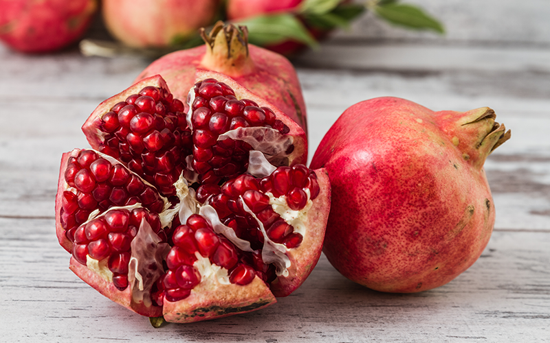 One pomegranate split down the middle sits in front of two whole pomegranates. The seeds of the open pomegranate look deep red, glistening, and delicious.