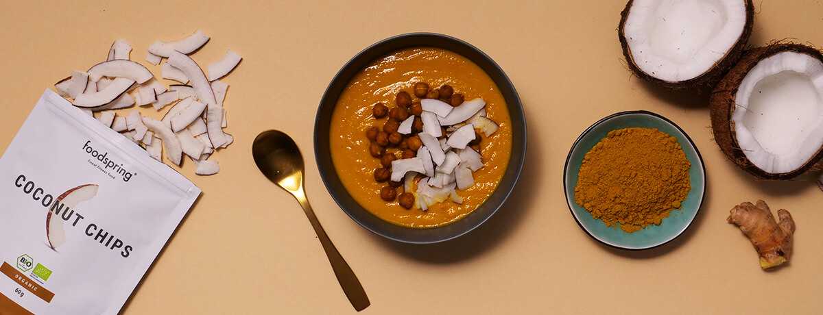 A bowl of carrot ginger soup topped with coconut chips and crispy chickpeas sits next to a halved coconut, a knob of ginger, and a teal bowl filled with a powdered medium-brown spice.
