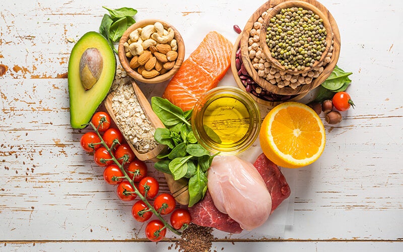 a photo of a selection of foods in a heart shape that are okay for clean eating - nuts, avocado, salmon, orange, cherry tomatoes, lentils, spinach leaves, spices, oats and a bowlful of oil.