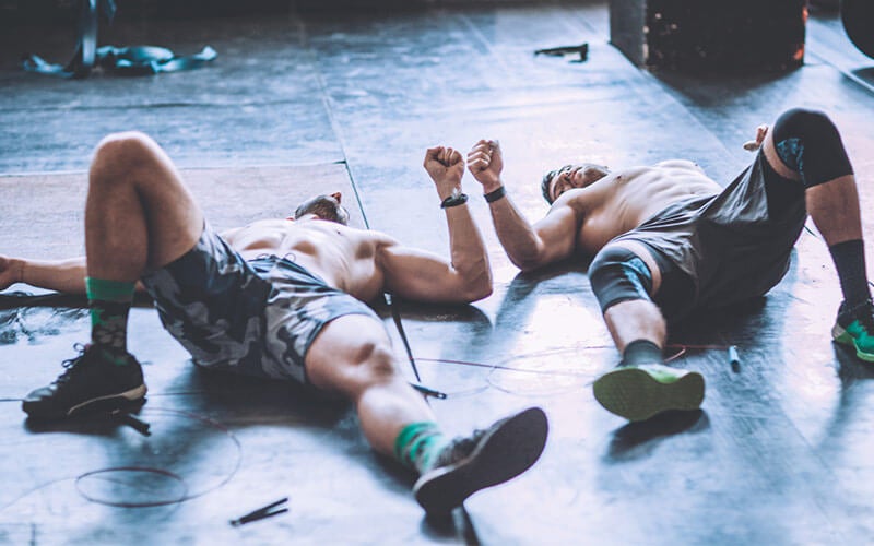 Two shirtless white men lie on the floor looking exhausted after a high-intensity workout