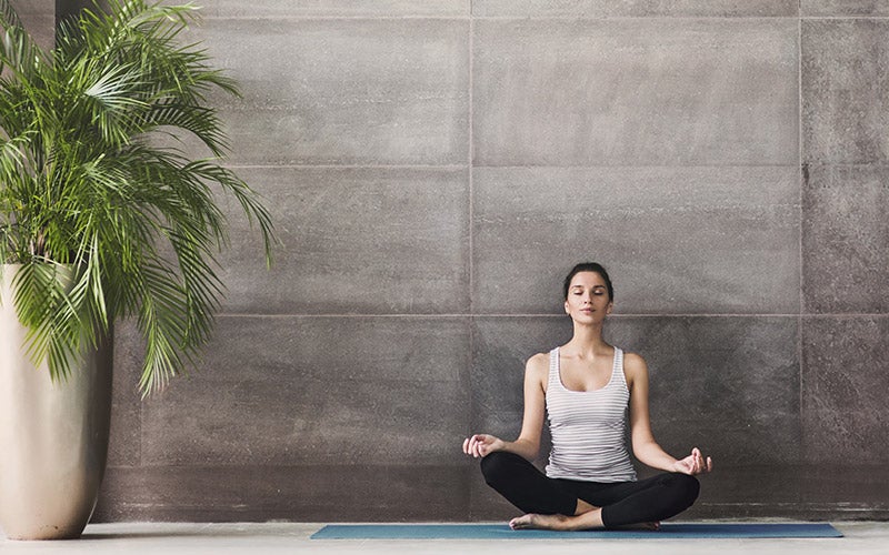 Wondering how to reduce stress? You might try meditation, as this white woman in a white tanktop shows. She sits cross-legged and closes her eyes, hands resting palms-up on her knees.