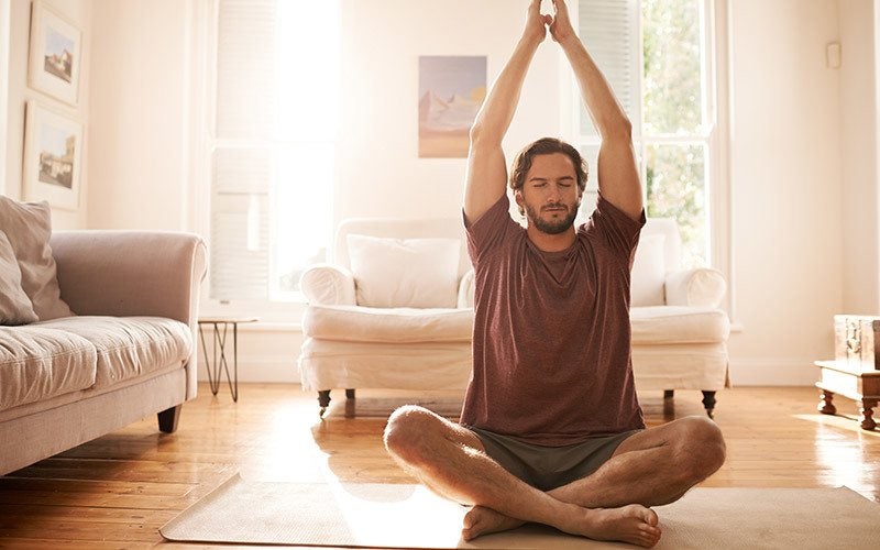 A white man sits with legs crossed on a yoga mat in a living room furnished with sofas. He wears a red t-shirt and stretches his arms over his head with his eyes closed.