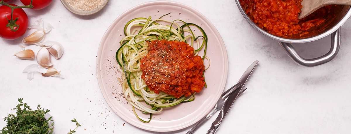 A white plate full of bright green and white zoodles (courgetti) with a vibrant red lentil-based bolognese.