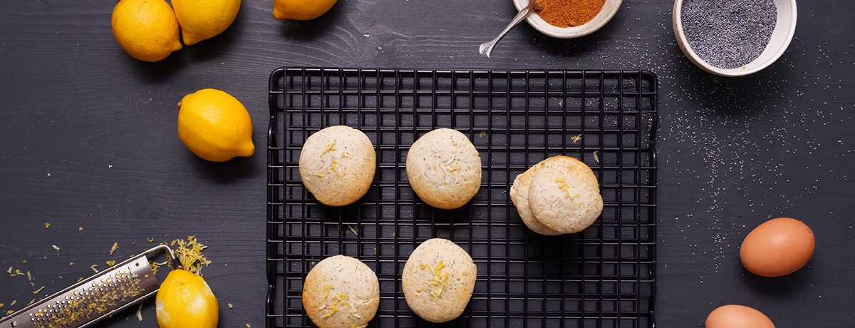 A black cooling rack holds four light golden lemon poppyseed cookies and a stack of three next to those. Surrounding the cooling rack are lemons, a zester, two eggs, a bowl of poppyseeds, and a bowl of brown sugar.