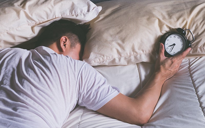 A white person with short black hair lies facedown in bed holding a black-and-white analog alarm clock. A good daily routine includes getting up right away when the alarm goes off!