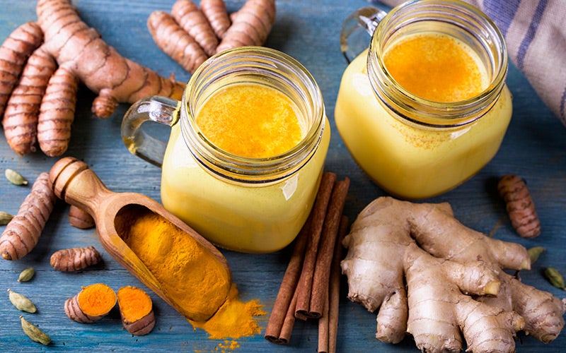 Two Mason jars full of golden yellow liquid surrounded by ginger and turmeric roots, a wooden scoop of turmeric powder, cardamom pods, and cinnamon sticks to make a golden milk. This is a great tip on how to boost your immune system.