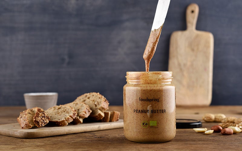 A jar of peanut butter sits next to slices of Protein Bread on a wooden cutting board, with peanut butter dripping from a metal knife above the jar, answering the question: are peanuts good for you?