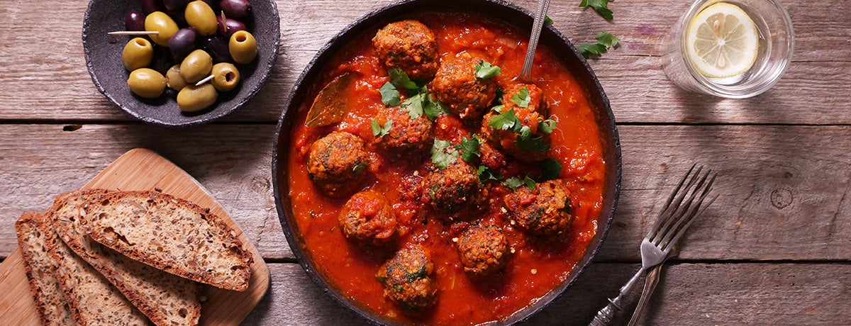 a dish of meatballs in tomato sauce with a bowl of olives, a glass of water with lemon slice, and a tray of bread slices on the side