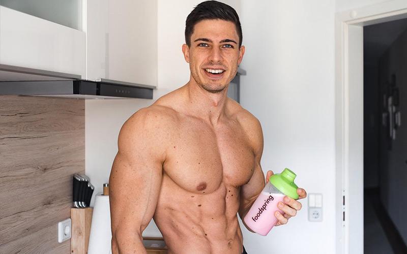 a photo of Julius Ise, a brown-haired, muscular white man, standing shirtless and holding a foodspring shaker with a pink shake inside.
