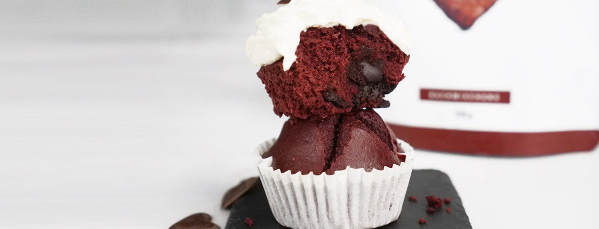 Two red velvet protein muffins stand, one stacked on top of the other. The top muffin is split open, revealing a fudgy center, and is topped with a bright white cream cheese frosting. The bottom muffin sits inside a white paper muffin cup. 