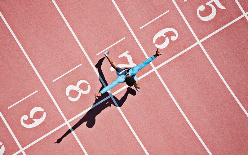 A view from above of an athlete of color with arms outstretched, left leg bent to the front and right leg extended behind them on a red track surface.