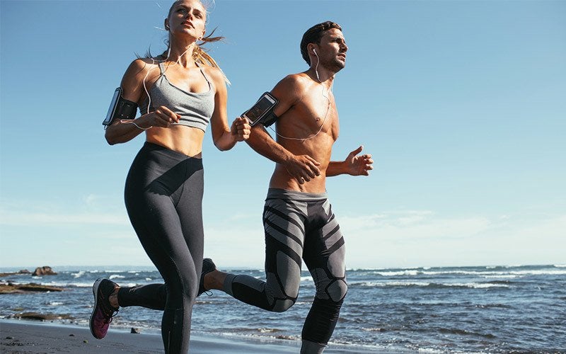 A white man and a white woman go jogging on a beach with a clear blue sky behind them.