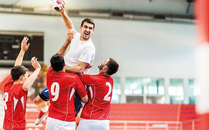 A grimacing white man jumps above opposing team members while playing handball, one of many types of sports to try out