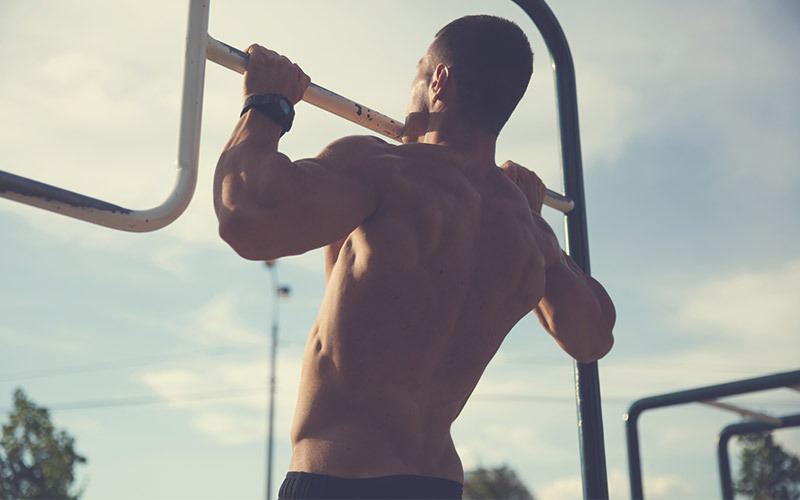 A shirtless white person with a flat chest is seen from behind doing a pull-up outdoors.