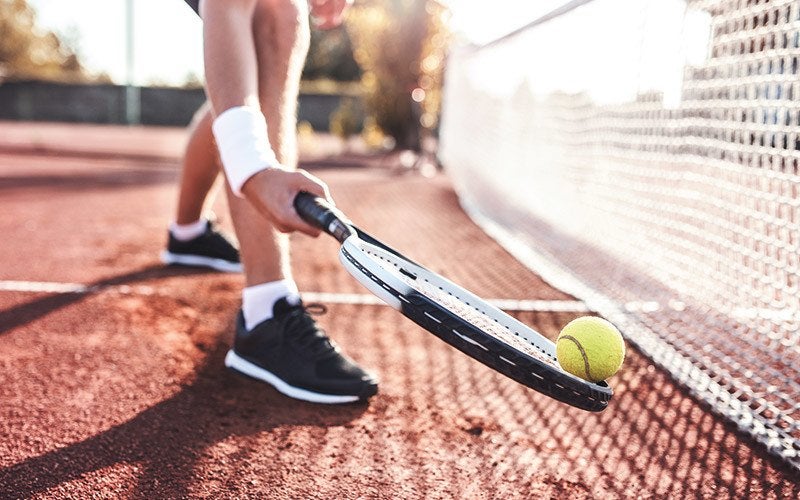 The leg and arm of an athlete are visible as they hold a tennis racquet to pick up a tennis ball from a red clay court with the net visible nearby.