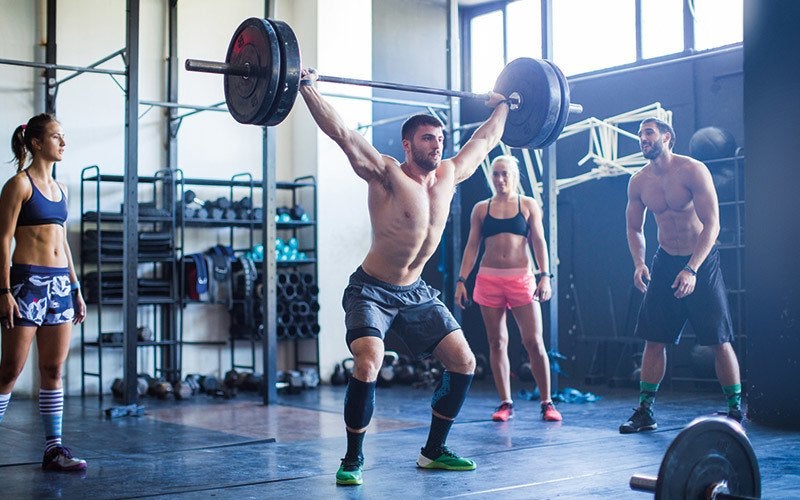 Two shirtless white men and two white women in sports bras inside a gym. One of the men lifts a weighted barbell above his head.