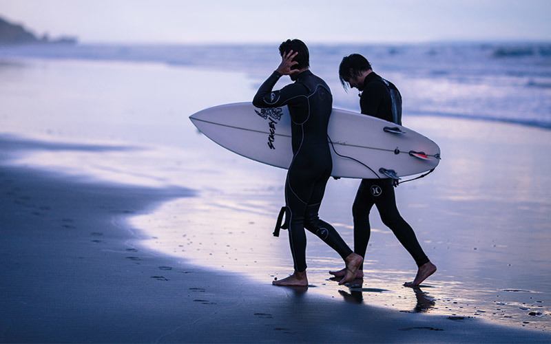 Two white men on a beach in long wetsuits carrying a surfboard. Surfing is one of many types of sports to choose from for outdoors enjoyment.