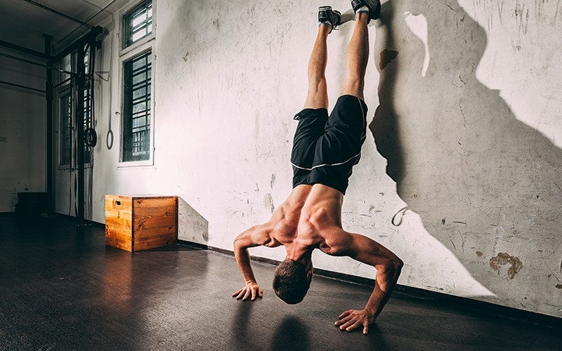 Handstand pushups are one way to get fit without equipment. Here a shirtless white man in black gym shorts demonstrates against a concrete wall.