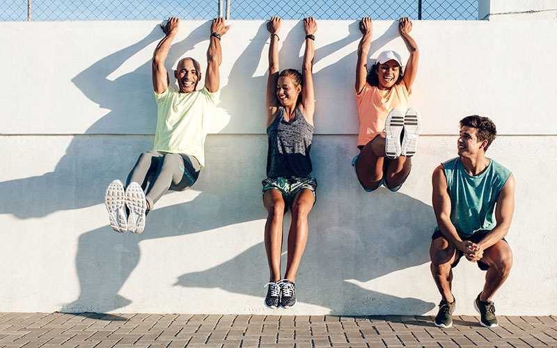 A group of two men and two women in sporty clothing. One man of color and two women of unclear skin color are using their fingertips to hang from the top of the wall behind them. The man squats next to the group and smiles at them.