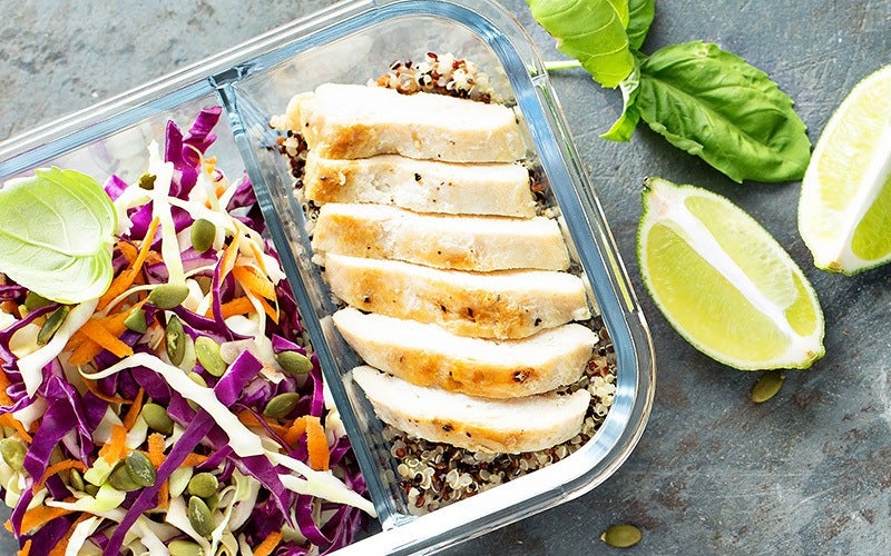 A glass food storage box holds a meal with quinoa topped with sliced chicken breast on one side of its divider while the other side has sprouts and shredded salad. On the side is lime quarters.