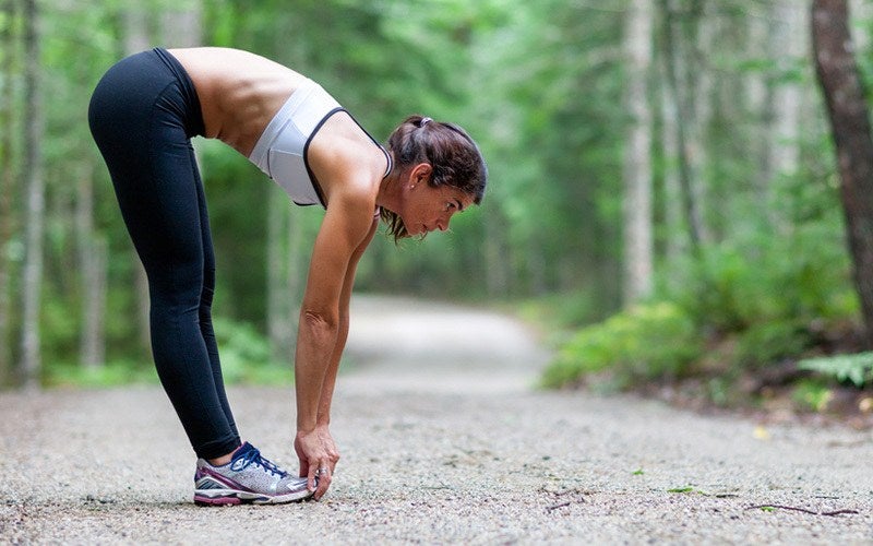 A woman of color reaches down to touch her toes while exercising outdoors on a forest path.