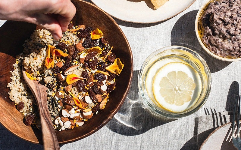 a white person's hand dips into a wooden bowl full of a quinoa-based salad with raisins, almonds and flower petals