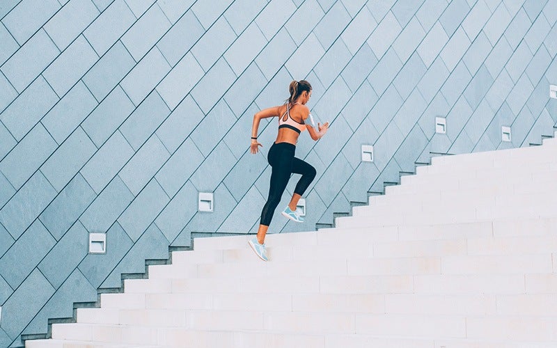 A white woman seen from behind wears exercise gear and climbs a set of white stairs with a blue-tiled wall in the background. Exercise is a great way to lose weight quickly and for the long term.