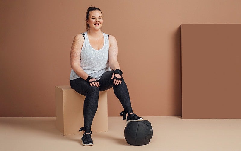 A fit white woman sits on a beige cube, wearing weightlifting gloves and resting one foot on a large slam ball. She is laughing.
