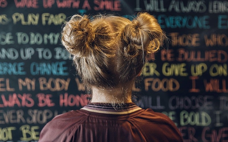 A white person seen from behind with two blonde buns looks at a vision board covered with colorful phrases. some of the visible words include play fair, everyone, change, never give up on, fuel my body, always be home, will choose, bold, never stop.