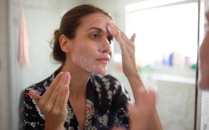A white woman applies a white foamy face mask in a mirror