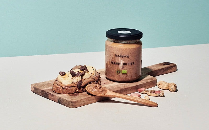 A jar of foodspring organic peanut butter sits on a cutting board next to a slice of bread gaily topped with a healthy portion of peanut butter, banana slices, and chocolate chunks