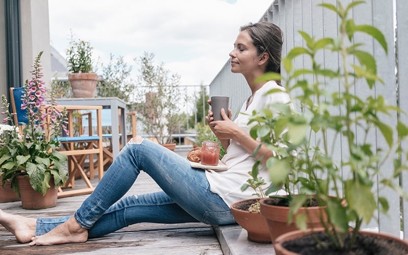 A white woman with light brown hair relaxes on a balcony, closing her eyes while cradling a gray mug and balancing a plate with a croissant and a pink jar of jam in her lap. She is clearly enjoying the relaxation she gets from intuitive eating.