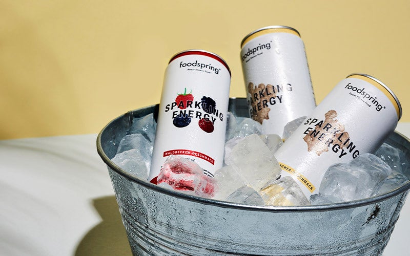 Three cans of Sparkling Energy Water - two ginger, one berry - sit in a metal tub full of ice cubes in front of a yellow background.
