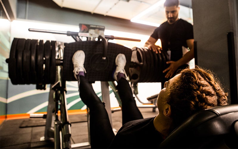 An athlete with French-braided hair reclines on a leg press machine as a bearded man spots her