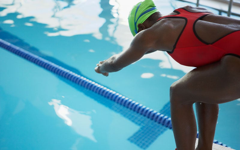 A woman of color in an orange swimsuit and bright yellow swim cap stands in the ready position before diving into the pool