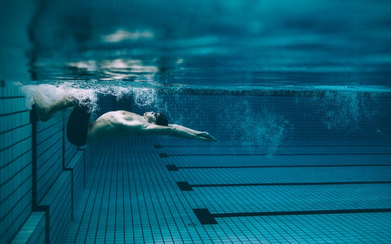 A topless white man in a swimming cap underwater pushes off from the wall of a pool. His arms are extended over his head while his knees are bent. He is on his back.