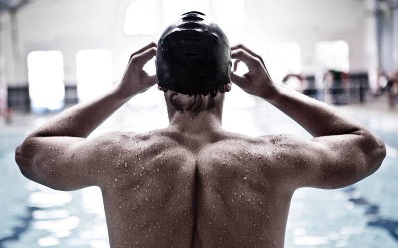 The top of a white topless swimmer's back, covered with droplets, wearing a black swim cap. The swimmer's hands are curved near the front of their face indicating they are adjusting their goggles.