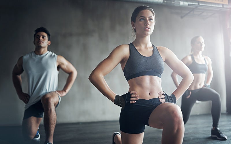Two women and a man do lunges in a gym to strengthen the sports habit. Check out our workouts page to get into your sports habit!
