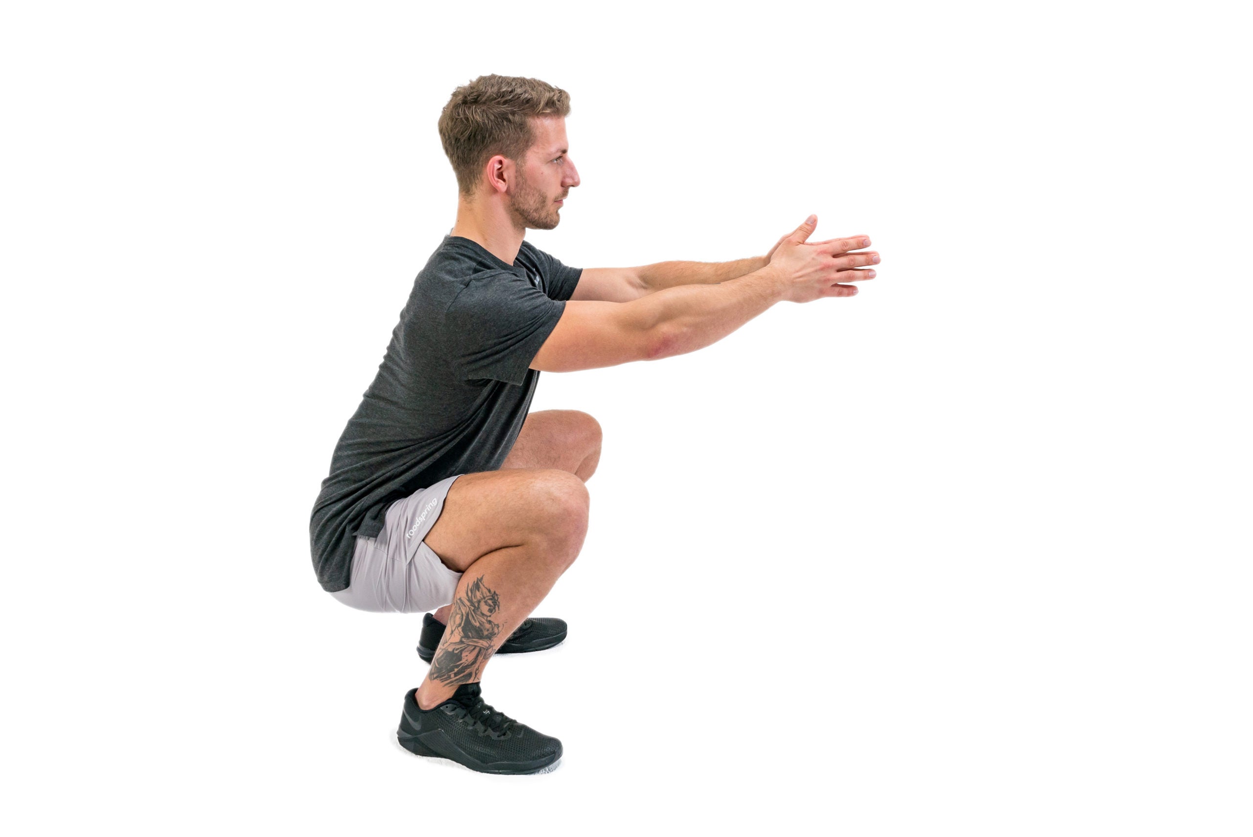 A white man in a black t-shirt and gray gym shorts demonstrates the appropriate ending position for an air squat. His arms are stretched in front of him, his knees fully bent and his calves touching his thighs. His back is straight and his rear end is as close to the ground as he can get it.