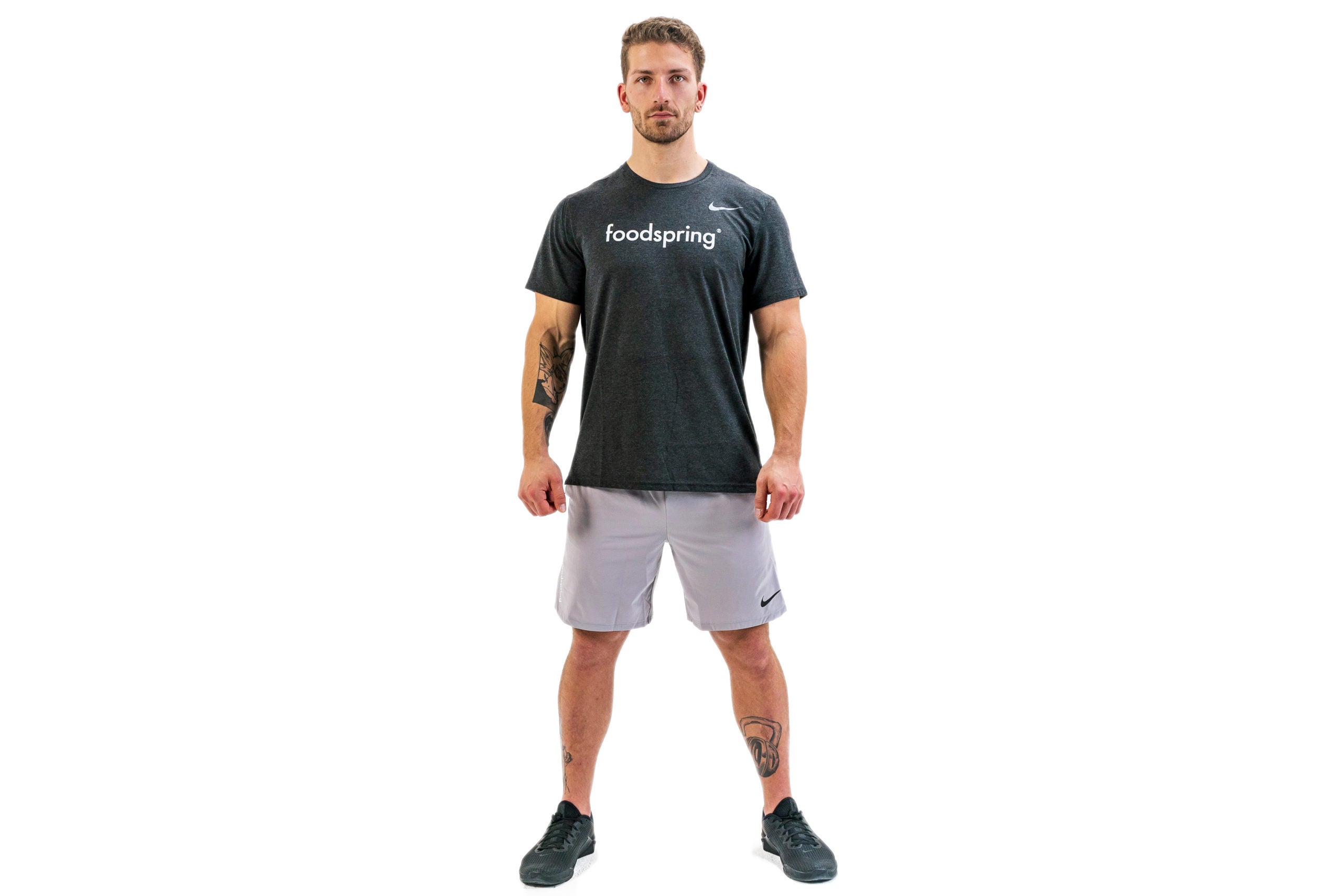 A tattooed white man in a black foodspring t-shirt and gray gym shorts shows the starting position for air squats. He is standing with his back straight, arms at his sides and feet shoulder-width apart.