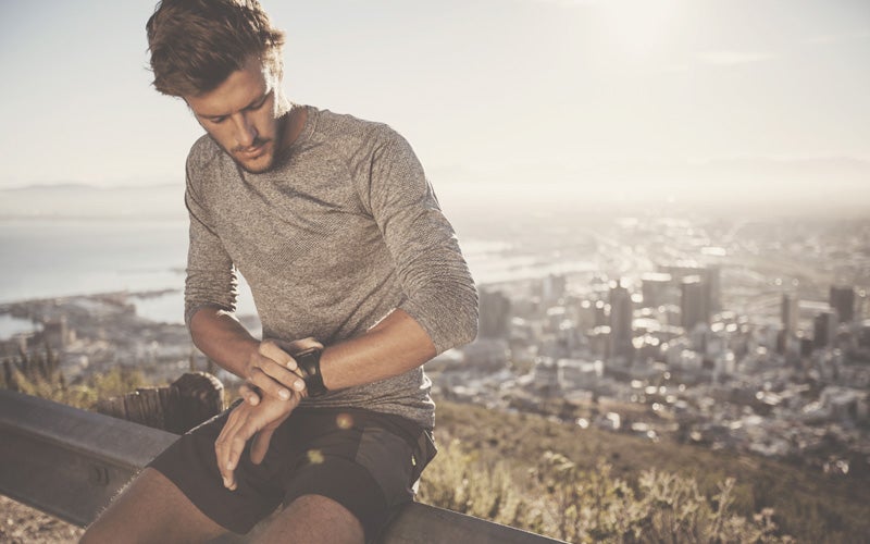 A white, short-haired man sits on a highway railing checks his pulse watch with a city gleaming in the distance behind him. How does exercise reduce stress? Maybe he knows.