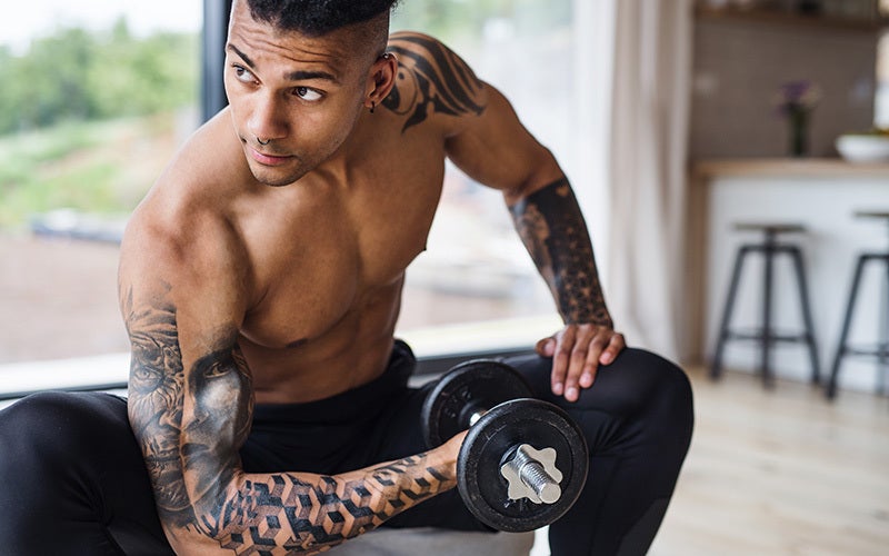 A shirtless, tattooed man of color does bicep curls at home in front of a glass door.